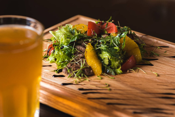 Fresh vegetable salad with pieces of meat on a wooden tray on a dark background next to them is a glass of light beer. - Photo, Image