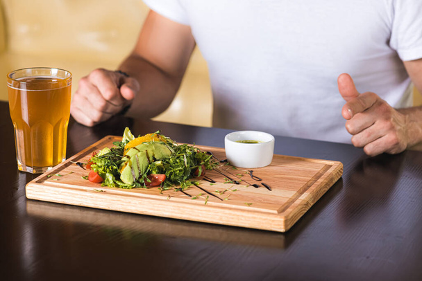 emotionaalinen nuori mies istuu pöydässä, odottaa syödä, hänen edessään vihannessalaatti, jossa on vihanneksia ja avokadoa ja lihaa. - Valokuva, kuva