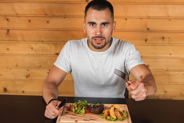 емоційний молодий чоловік сидить за столом, чекаючи їжі, перед нею стейк з картоплею фрі і салат з зеленої
 - Фото, зображення