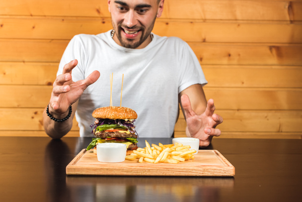 młody, przystojny emocjonalny mężczyzna w białym T-shircie siedzący przy stole przed nim drewniana deska na której pyszny hamburger, przestrzeń układu dla SMS-a lub projektu - Zdjęcie, obraz