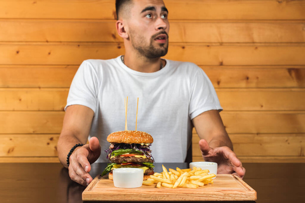 jonge, knappe emotionele man in een wit T-shirt zittend aan een tafel voor hem een houten bord waarop een heerlijke hamburger, lay-out ruimte voor een sms of design - Foto, afbeelding