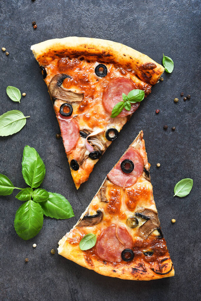 Plasterki pizzy z salami, grzybami, oliwkami i bazylią na czarnym tle. Widok z góry. - Zdjęcie, obraz