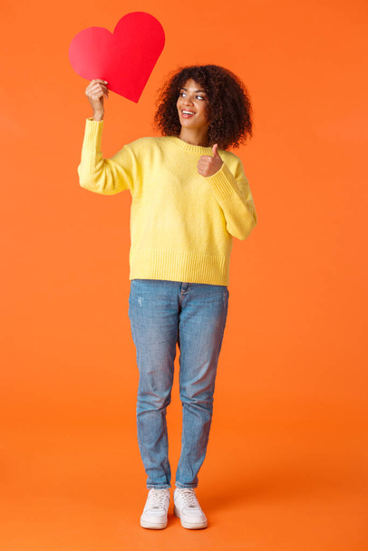 Полноразмерный вертикальный выстрел удовлетворен и доволен, красивый африканский хипстер девушка чувствует себя хорошо без отношений, держа ее красный знак сердца и дать большой палец вверх, оранжевый фон
 - Фото, изображение