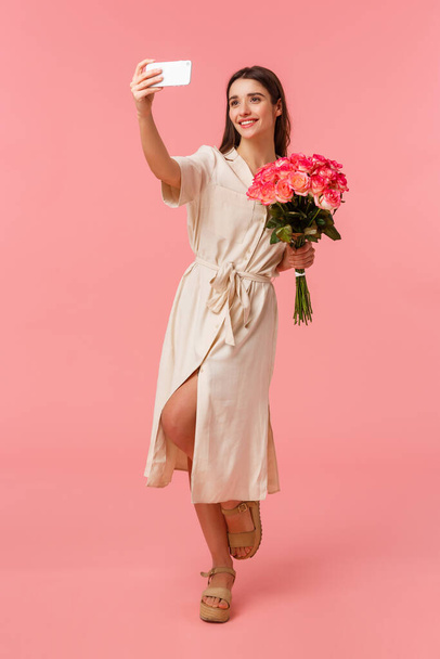 In voller Länge vertikales Porträt elegant gut aussehende Geburtstagskind in Kleid, Selfie mit Blumen, erhalten schöne Strauß Rosen, lächelnd und fotografierend, um online zu posten, rosa Hintergrund - Foto, Bild