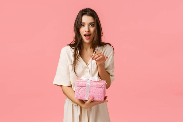 Excitement, tenderness and celebration gesture. Feminine surprised and excited alluring brunette girl in dress, wrapping gift, look amused and wondered, holding cute present box, pink background - Photo, Image