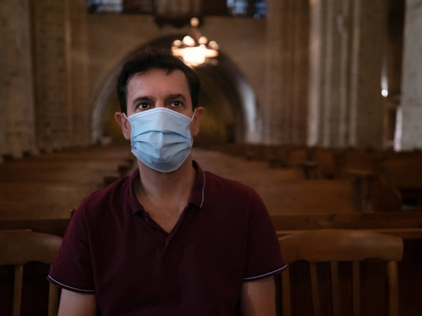 Горизонтальний портрет чоловіка в хірургічній масці, що сидить і молиться в церкві
 - Фото, зображення
