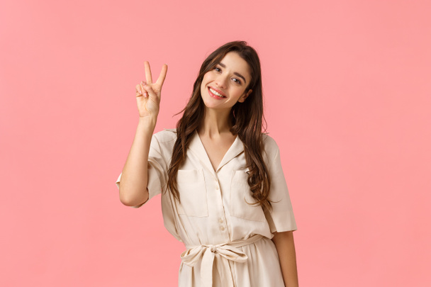 Women, beauty and advertising concept. Cheerful tender brunette woman feeling relaxed and delighted, smiling lovely, tilt head coquettish as wearing dress showing peace sign, pink background - Photo, Image