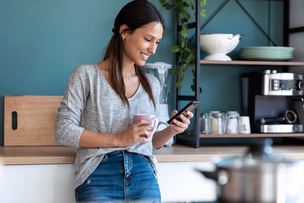 Laukaus hymyilevä nuori nainen käyttää matkapuhelintaan juodessaan kupin kahvia keittiössä kotona. - Valokuva, kuva