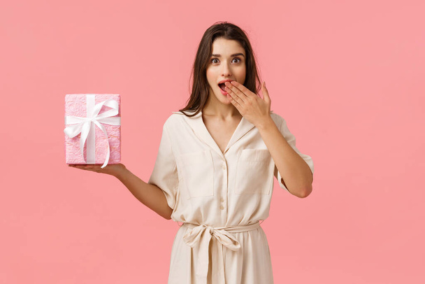 Surprises, holidays and celebration concept. Amused and cheerful attractive young brunette girl in dress, gasping open mouth amazed, receive wonderful birthday gift, pink background - Photo, Image