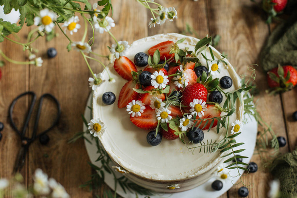 Σε ένα τραπέζι ανάμεσα στα καλοκαιρινά χόρτα, ένα κέικ είναι διακοσμημένο με μούρα και λουλούδια. - Φωτογραφία, εικόνα