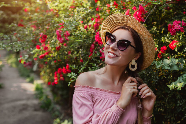 Lächelnde Frau mit Strohhut-Sonnenbrille spaziert an blühenden Rosen im Sommergarten vorbei. Mädchen genießt Blumen im Park. Sommer-Outfit. Modische Accessoires - Foto, Bild
