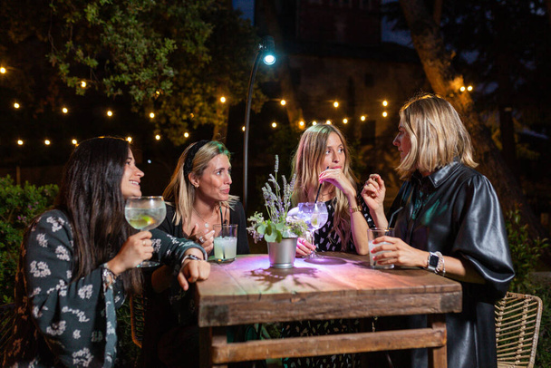 4 chicas hablan en la terraza de un club (pub) por la noche, se visten de negro, y tienen vasos de cóctel y tallos. Son caucásicos, 3 rubias y una morena. sobre una mesa de madera alta
. - Foto, imagen