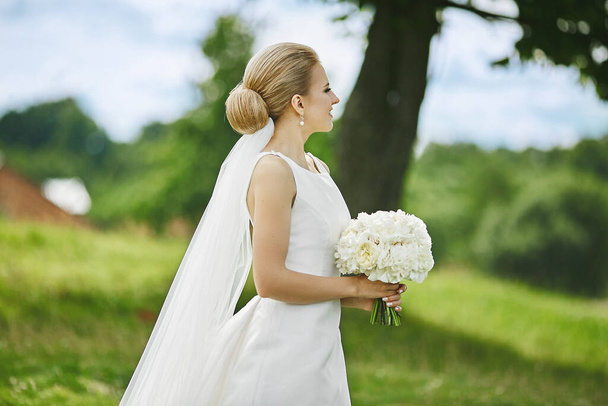 Beautiful and elegant young blonde woman in stylish wedding dress with bouquet of white flowers in her hands posing outdoors - Φωτογραφία, εικόνα