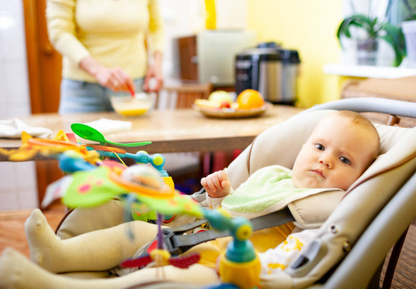 Blurred charming smiling mom sits at the table and rejoices at her little cheerful newborn baby girl playing in her baby seat with toys. Happy motherhood concept - 写真・画像