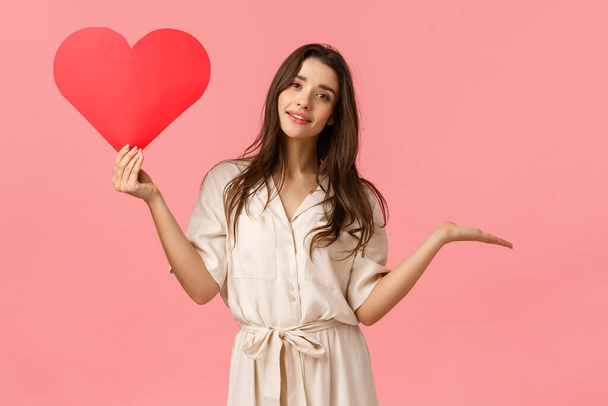 Surprised girl searching true love. Amused and carefree cute young woman brunette, raising hand indecisive and unsure smiling, holding valentines heart card, standing pink background - Foto, immagini
