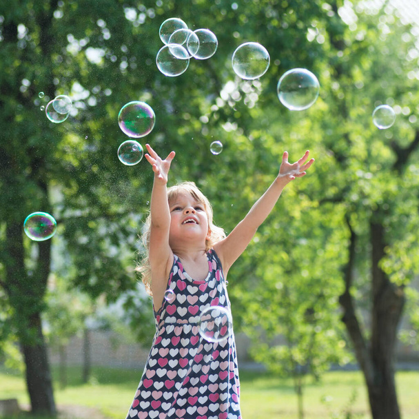 Милая маленькая девочка в платье играет с мыльными пузырями на открытом воздухе
. - Фото, изображение
