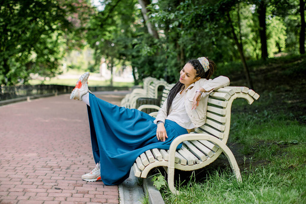 Outdoors portrait of modern hipster girl with dreadlocks wearing blue skirt and white t-shirt, sitting on the bench in a city park with one leg raised and having fun - Foto, Bild
