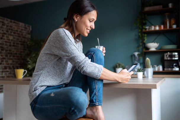 Laukaus hymyilevä nuori nainen käyttää matkapuhelintaan syödessään jogurttia keittiössä kotona. - Valokuva, kuva