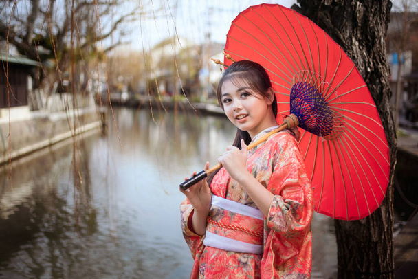 Ασιάτισσες τουρίστες. Γιαπωνέζα που φοράει κιμονό κρατώντας κόκκινη ομπρέλα. Όμορφη γυναίκα φορώντας παραδοσιακά ιαπωνικά κιμονό Σε διάσημα τουριστικά σημεία στον ποταμό Yanagawa Fukuoka, Ιαπωνία. - Φωτογραφία, εικόνα