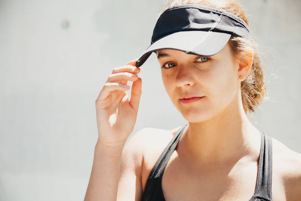 Portrait of a beautiful sporty young woman with stylish sportswear and a visor cap posing against a white wall in the street in a hot day - Foto, imagen