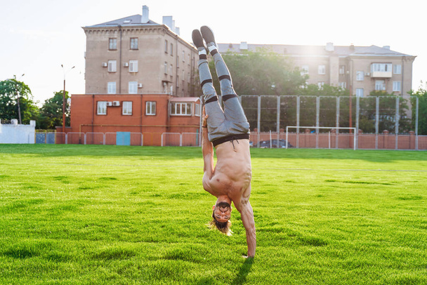 Standing on one hand upside down. Strong, muscular inflated extreme man. Young athlete of Caucasian nationality standing on the football field and doing different physical exercises. - Photo, Image