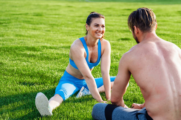 Beautiful, muscular, pumped-up athletes sitting on grass on the football field opposite each other, doing morning exercises. Smiling, enjoying the sun. Exercises in a pair, motivation for each other. - Valokuva, kuva