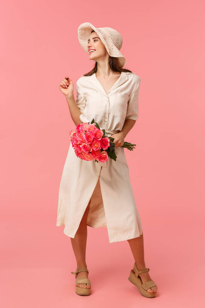 Full-length vertical portrait tender and romantic happy young woman smiling wearing dress and hat, turning left holding roses, receive bouquet flowers, standing pink background - Фото, изображение