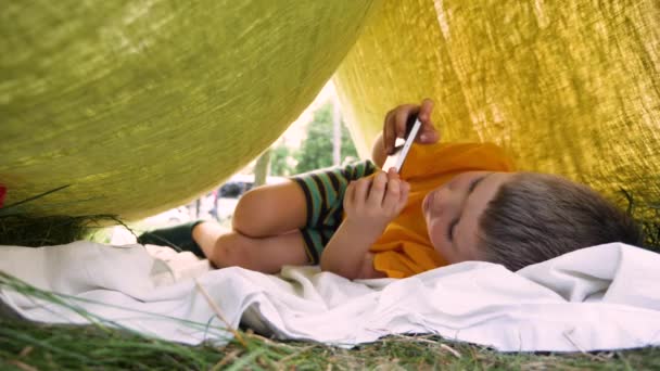 Trekking kis ház gyerekeknek. Egy srác telefonnal, aki videojátékozik. Fiú töltik nyári sátorban mobil apps. - Felvétel, videó