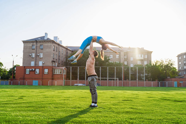 Zdolna młoda para uprawiająca akro jogę w parku lub na stadionie polowym. Mężczyzna stojący na trawie i balansujący swoją kobietą na rękach. Szczęśliwi, sportowi młodzi sportowcy. - Zdjęcie, obraz