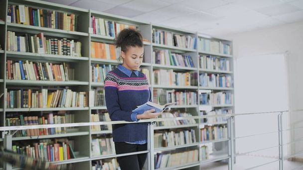 Porträt der jungen, lockigen Afroamerikanerin, die das alte Buch liest, während sie neben den Bücherregalen in der Bibliothek steht. - Foto, Bild