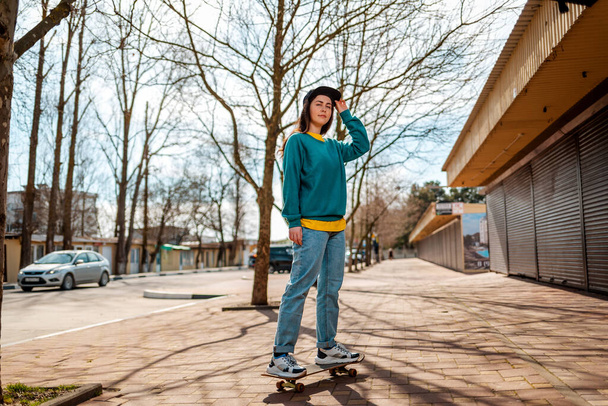 A young Caucasian hipster woman poses standing on a skateboard. In the background, an alley. Concept of sports lifestyle and street culture. - Photo, Image