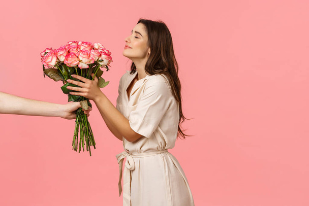 Romance, valentines day and happiness concept. Gorgeous young woman receiving delivery, smelling pretty roses as hand extending bouquet to girl, smiling delighted, got surprise gift, pink background - Photo, Image
