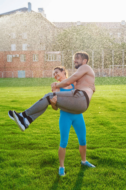 Funny photo. Cute couple of sportsmen having fun during sports training. Woman raised huge pumped up man and holding him in her hands. Laughing together. - Фото, зображення