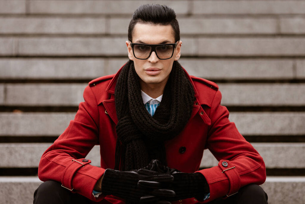 LGBTQ community lifestyle concept. Young homosexual man sits on a stairs. Handsome fashionable gay male model poses in cityscape outdoors. Wears red coat, gloves, sunglasses and black scarf - Фото, зображення