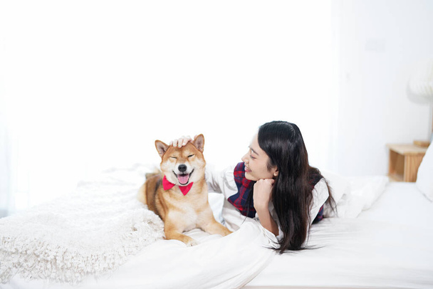Háziállat-barát koncepció. Egy ázsiai nő játszik egy kutyával egy fehér ágyon a hálószobában.. - Fotó, kép