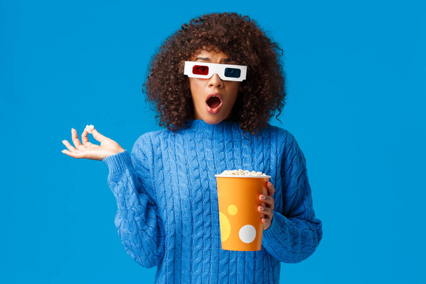 Zszokowany i zaskoczony młody słodkie afrykańsko-amerykańska hipsterka dziewczyna ogląda film akcji, dramat dysząc i spadek szczęki jak jedzenie popcornu i zobaczyć szokującą scenę w filmie w okularach 3D, niebieskie tło - Zdjęcie, obraz