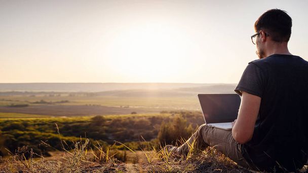 ノートパソコンを持った男が谷の素晴らしい景色を眺めながら山の端に座っている。セルラーネットワークのブランドカバーの概念。5G. - 写真・画像