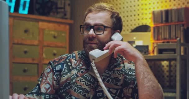 Běloch v brýlích a barevné košili sedí ve špinavé místnosti osmdesátých let. Muž mluví na domácím telefonu s drátem a pracuje na počítači v 90. letech. Retro styl na volné noze. - Záběry, video