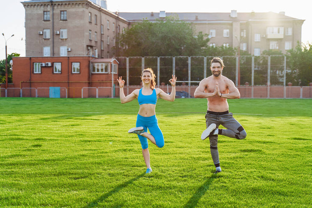 Pair of young athletes of Caucasian nationality standing on the football field and doing different physical exercises. Standing with arms complex on their chests and a raised, bent leg. Laughing. - Photo, Image