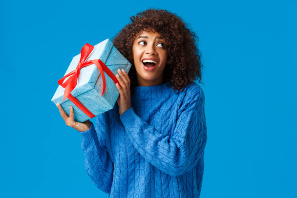 期待、休日や冬の概念。陽気なアフリカ系アメリカ人の女性がプレゼントで箱を揺らし、アンラッププレゼントをしたい好奇心と面白がって、笑顔の夢の中に何を参照してください - 写真・画像
