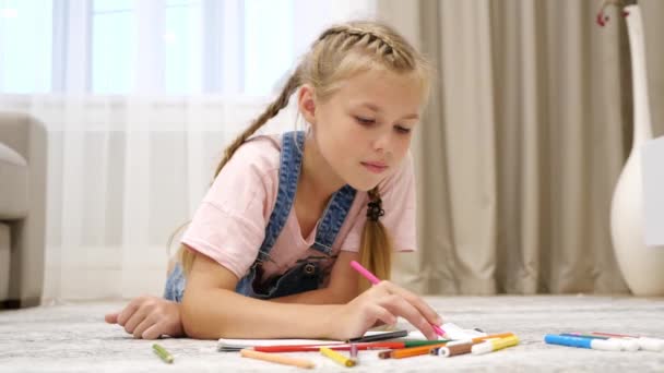 Cute preteen girl lying on floor and drawing on paper in living room - Footage, Video