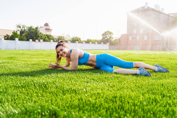 Girl in blue fitness clothes lying on grass stadium. Brunette advertises green lawn on football field. Woman has sporty lifestyle. Rest after training at city stadium. Model posing. - Photo, Image