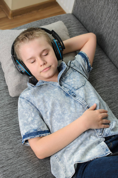 Niño adolescente con auriculares escuchar música en el sofá en casa. Adolescente disfrutar de la música - retrato de niño rubio mientras está acostado en el sofá en el interior. Tiempo de relajación. - Foto, imagen