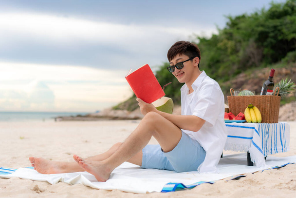 Nuori aasialainen mies käyttää aurinkolaseja rentouttaa lukemalla kirjaa valkoisella hiekkarannalla ja lähellä merta trooppisilla hedelmillä taustalla. Kesä, loma, loma ja onnelliset ihmiset Thaimaassa käsite. - Valokuva, kuva