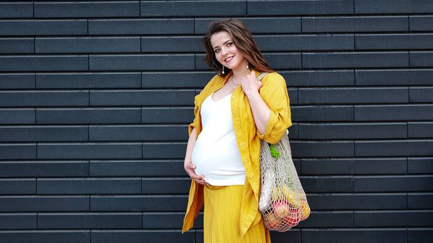Вагітна жінка в жовтому одязі з екологічною сумкою з фруктами та овочами на фоні чорної стіни. Безпластикова концепція Сталий спосіб життя
. - Фото, зображення