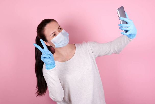 Portrait of a brunette girl in a medical mask and blue rubber gloves makes selfie with cellphone. - Фото, зображення