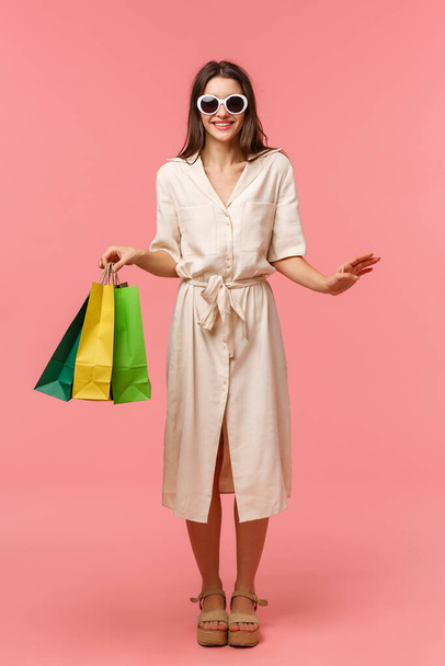 Full-length verticaal portret elegante goed uitziende vrouw op vakantie, winkelen en plezier hebben in winkelcentra, het dragen van een zonnebril, lichte jurk, draagtassen uit de winkel, staande roze achtergrond glimlachen - Foto, afbeelding