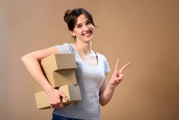 Un mensajero con cajas en la mano derecha muestra una señal de victoria con una sonrisa en las manos sobre un fondo beige
. - Foto, Imagen