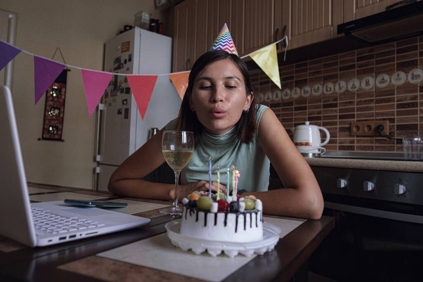 Mujer soplando la vela en el pastel de cumpleaños y haciendo videollamada. Chica celebrando cumpleaños en línea en tiempo de cuarentena
. - Foto, Imagen