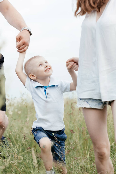 Ένα μικρό αγόρι να χαμογελάει και να κρατάει τα χέρια των γονιών του ενώ περπατούν στη φύση. Ευτυχισμένη οικογένεια - Φωτογραφία, εικόνα